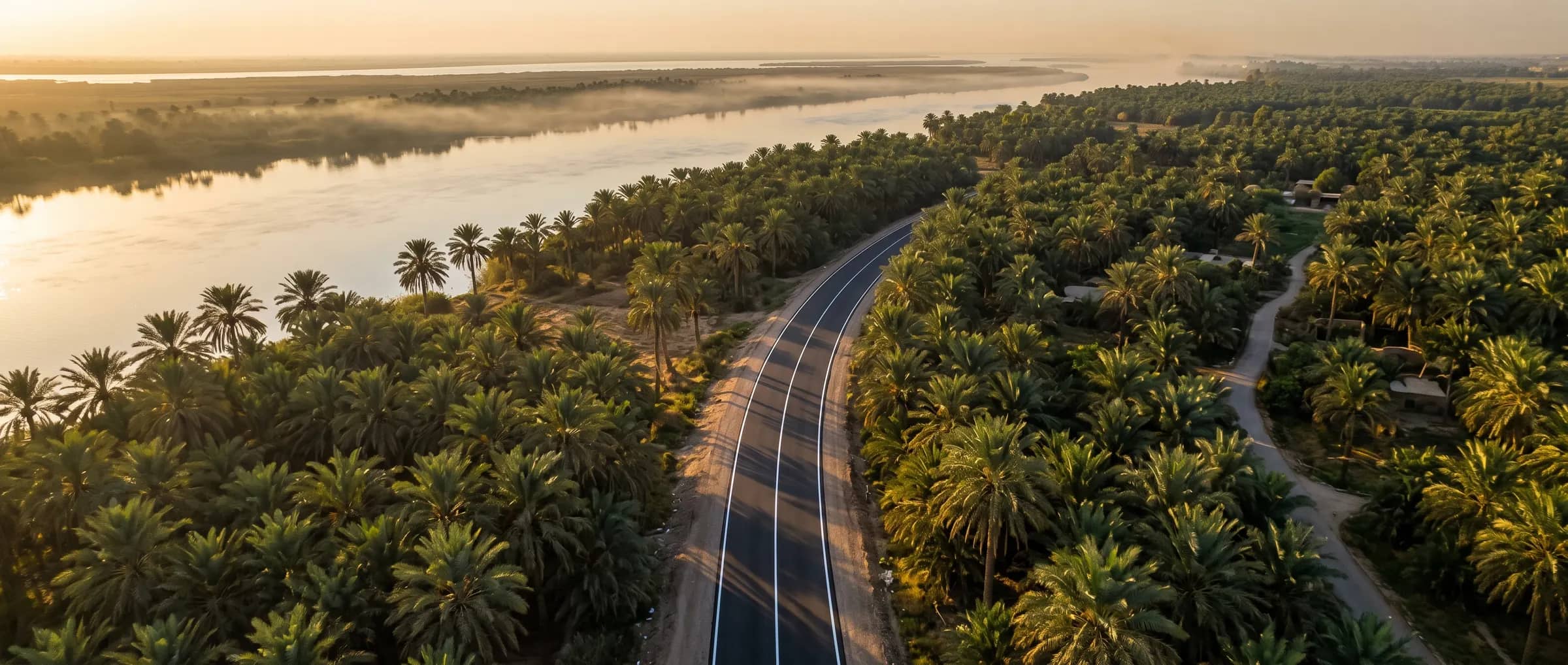 An aerial view at sunrise of a paved road with white lines running through palm groves beside a river